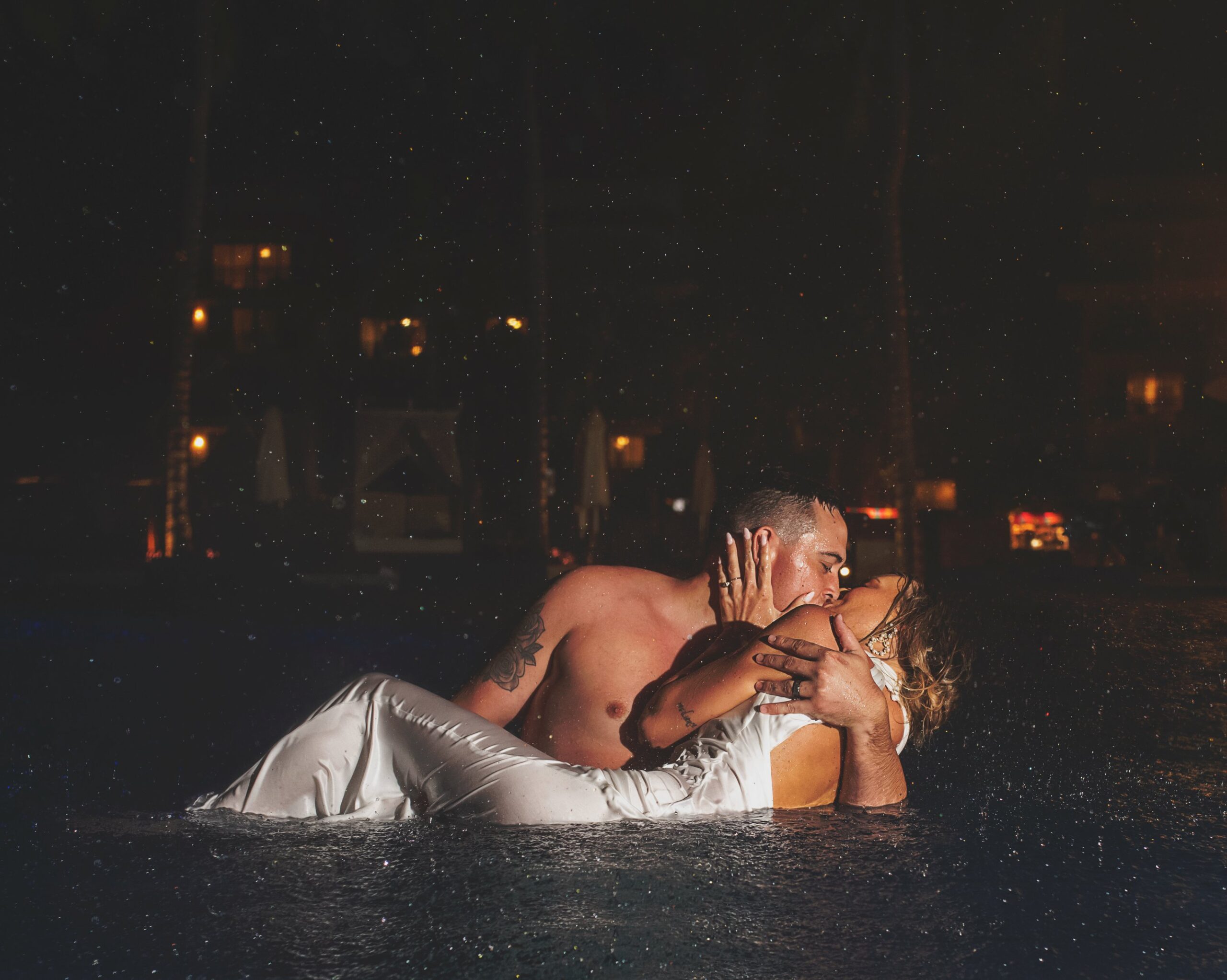 A bride and groom passionately kissing in a pool at night, both soaking wet after jumping in fully clothed, with raindrops visible around them.